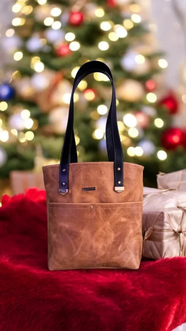 Brown leather tote bag with black handles in front of a decorated Christmas tree.